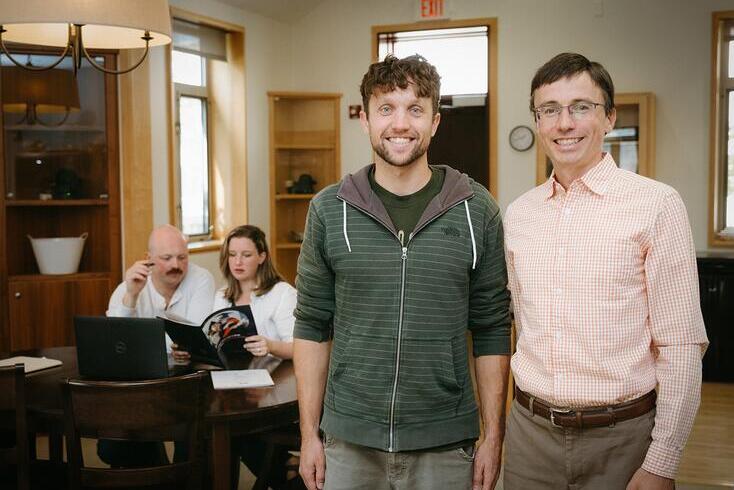 Two men are standing in a living room setting, smiling and looking into the camera. In the background, a man and a woman are sitting at a table reading a brochure.