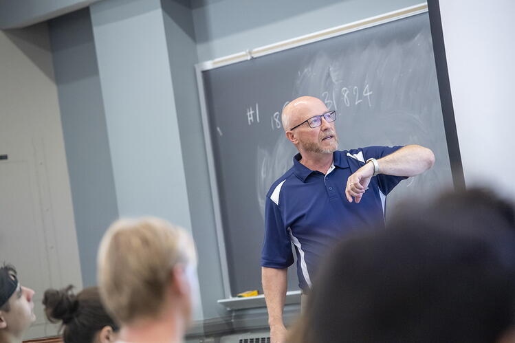 A man in a dark blue shirt and glasses gestures at a screen while teaching in a college classroom. Backs of students' heads are visible in the foreground.