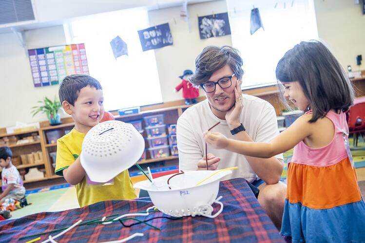 Two children play with white plastic colanders and pipe cleaners in a colorful classroom. A male teacher with glasses sits between them.