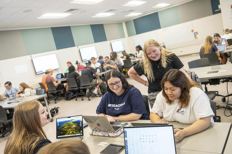 Five ALEC students collaborate around laptops in a classroom while other students work together in groups in the background.