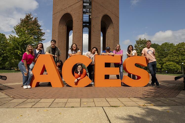 Students stand around giant ACES letters in front of the bell tower. 