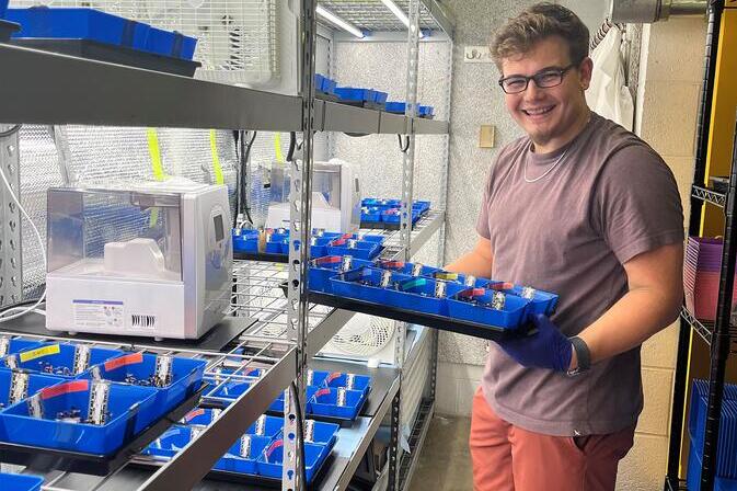 Liam Reynolds is holding a tray with blue containers next to metal shelves with similar trays.