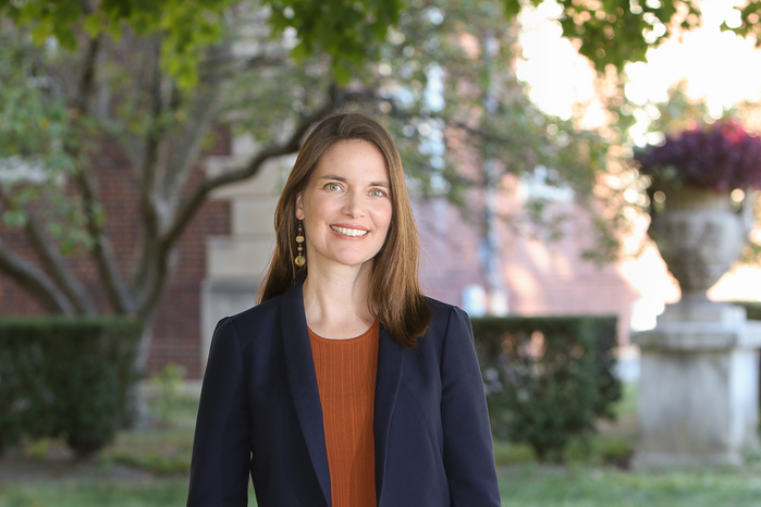 A smiling Sarah Low wears an orange shirt and blue blazer, standing outdoors on campus.