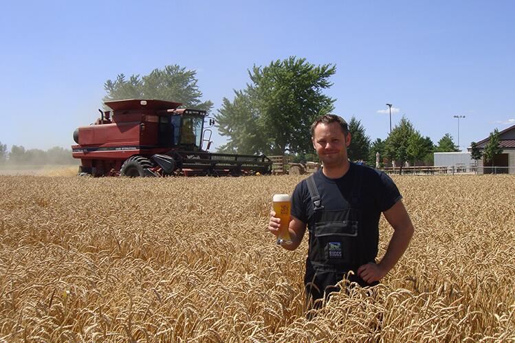 Matt Riggs stands in a field of wheat holding a glass of beer in front of a combine. 