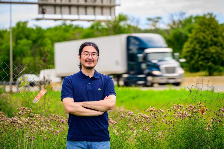 	 A man with glasses and a ponytail stands with arms crossed near a fence by a highway, with a blurred semi-truck passing in the background.