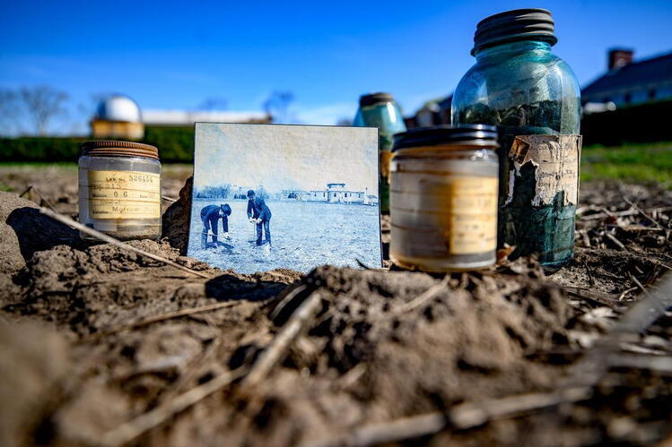 Federally funded agricultural research has been a cornerstone of the College of ACES since its inception. Photo: Fred Zwicky. A historic black and white photo showing two men in suits and hats shoveling soil in a field is propped against a set of three ancient-looking bottles holding old soil samples. The objects sit in an agricultural field