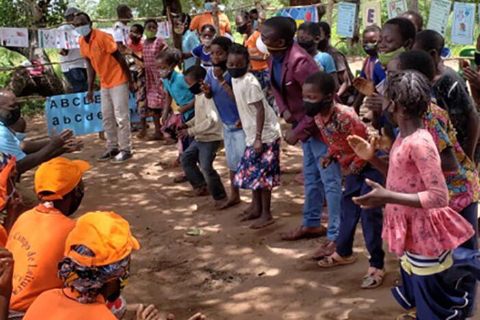 Children participating in out-of-school reading activities in Mozambique. A large group of African school children playing a game in an outdoor setting, with reading materials in the background