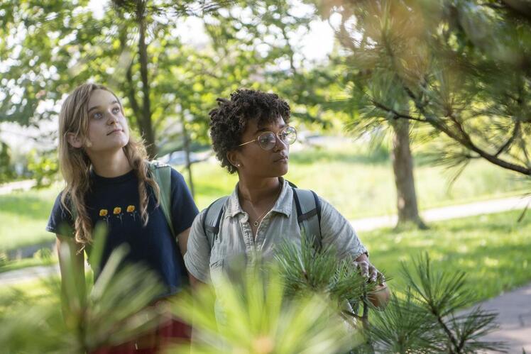 Two people stand outdoors under trees, looking intently at their surroundings. One wears a dark t-shirt, while the other has glasses and a light shirt with a backpack.