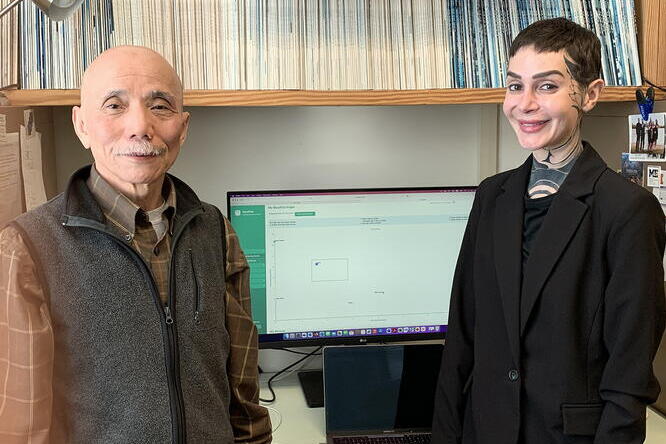  A man wearing a shirt and a vest and a woman wearing a black jacket are standing in an office setting in front of a computer and a bookshelf. 