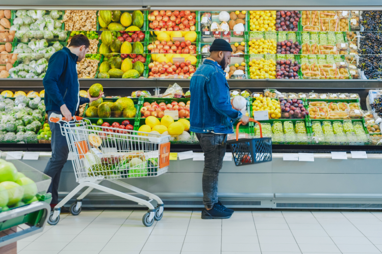 Two men choose fruits in the produce section of a grocery store. The background is filled with brightly colored fruits and vegetables.