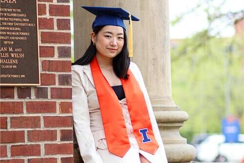 Nicole Park sits on a ledge wearing a white dress, cap, and stole. 