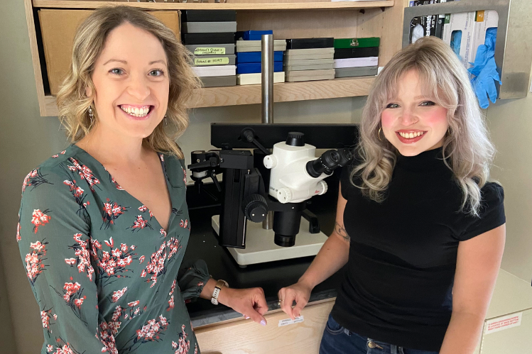 Two women smiling and standing beside a microscope in a lab, one wearing a floral shirt and the other a black shirt, with various equipment and books in the background.