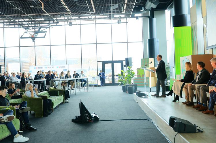Dean Germán Bollero stands at a podium in a bright, modern event space with large windows and Chicago-themed signage.