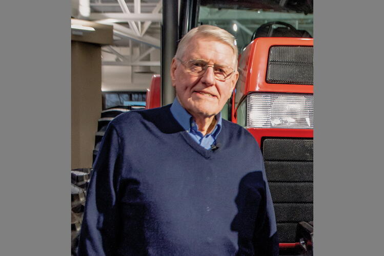 Paul Wallem. Photo courtesy of Farm Equipment magazine. A man with grey hair and glasses, wearing a blue sweater, stands in front of large agricultural machinery in a hall