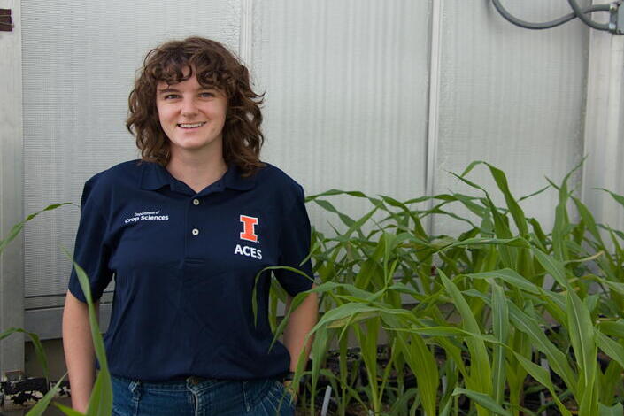 A woman with brown, curly hair smiles in front of plants in a greenhouse