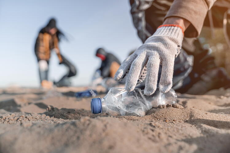 A gloved hand is picking up a plastic bottle on a beach, with other people collecting waste in the background
