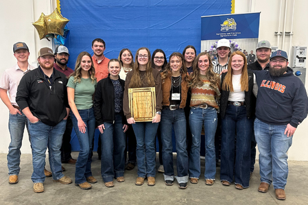 The U. of I. group at the SASES Spring Meeting, with their first place soils team plaque. Greta Hall is pictured in the front row wearing a green shirt. Sixteen young adults stand in a group. The woman at center holds a tan-colored plaque. Behind the group, a blue backdrop and gold balloons are visible.