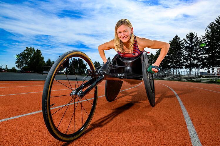 A person smiles while using a racing wheelchair on a track. A clear blue sky and trees are in the background.