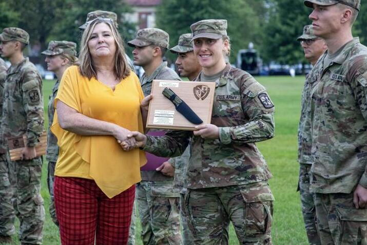 People in army fatigues stand at attention. In the center, a person in army fatigues holds a plaque and shakes the hand of a person in civilian clothing