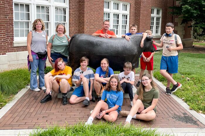 Nine students and three adults stand beside a bronze statue of a bull outside of the Stock Pavilion building. 