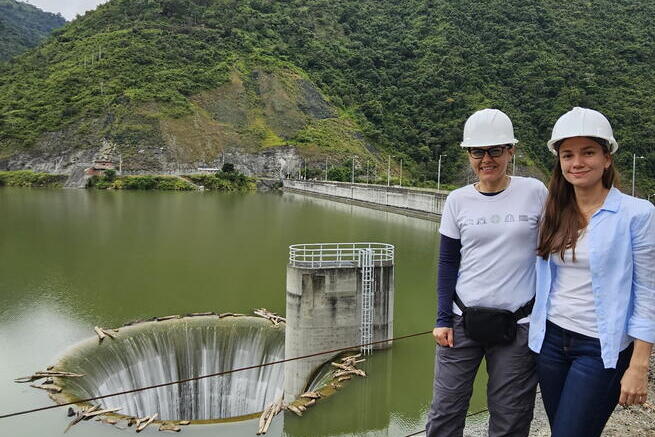 Two women wearing hard hats stand near a reservoir with a visible morning glory spillway and surrounding green hills in the background.