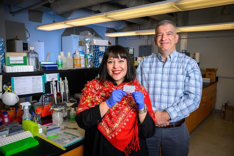 Two people stand in a laboratory; the woman in the foreground holds up a small object with her gloved hands while the man in the background looks on. Scientific equipment surrounds them.