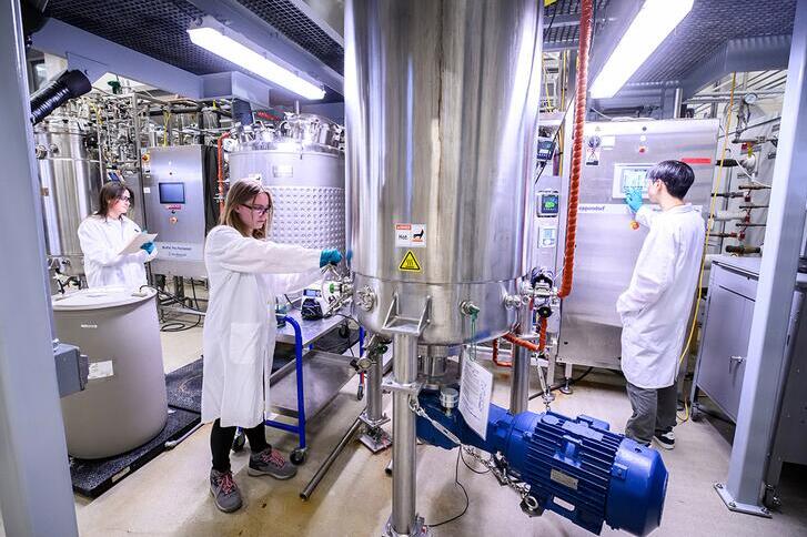 Three people in lab coats check instruments in a room filled with chrome tanks and lab equipment.