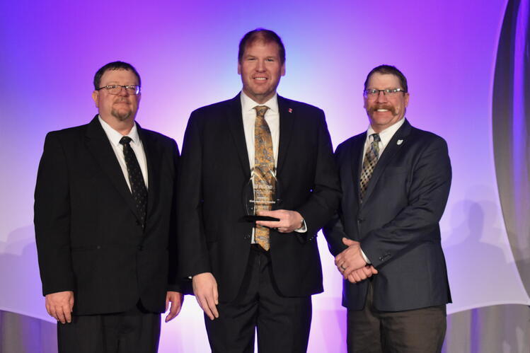 Three men in suits stand against a purple background. The man in the center is holding an award. 