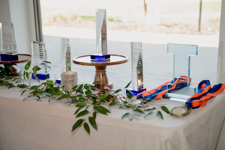 Orange and blue medals, glass awards, and a University Block I are placed on a table at the College of ACES Alumni Award Gala