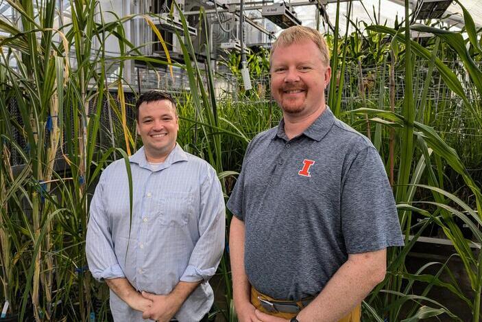 Two men stand in front of large plants in a greenhouse