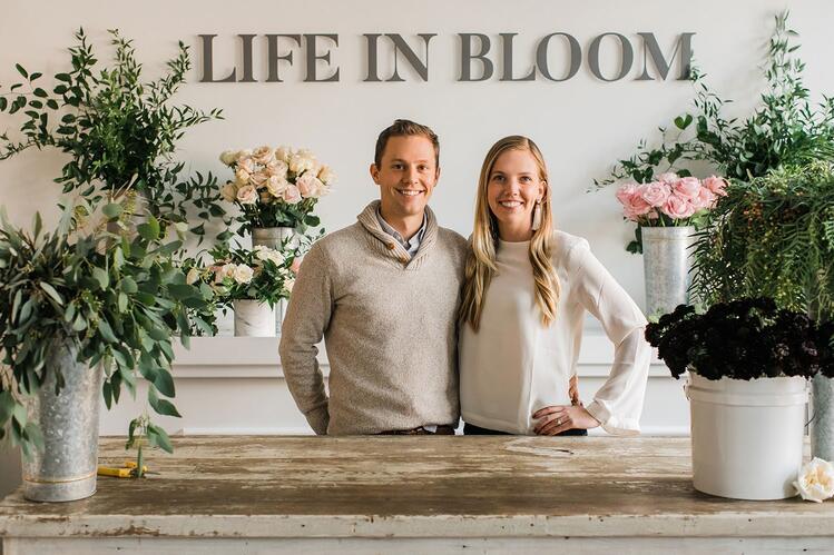 Tim Jung and Rachel Wyffels pose in front of a “Life in Bloom” sign, surrounded by flower arrangements.