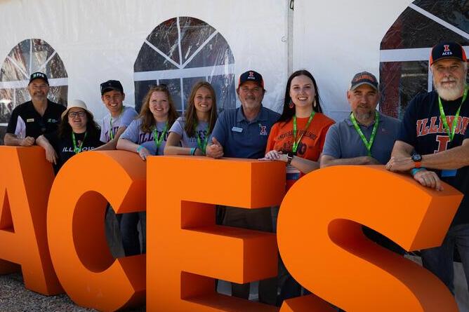 Media professionals and students pose behind a large orange ACES sign at the Farm Progress Show