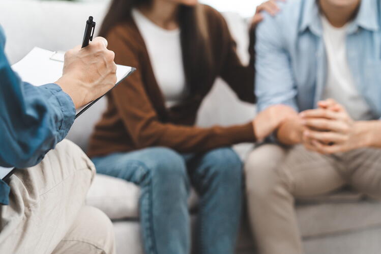 Better understanding of relationship development can help couples overcome challenges. A man and a woman, without their faces showing, sitting in a sofa holding hands, while the hand of another man is visible taking notes.