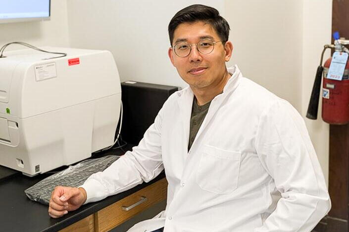 A man in a white lab coat sitting on a chair next to laboratory equipment. 