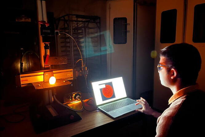 A man sits a a table ith a laptop computer in a dark room where a strong light is shining on an egg