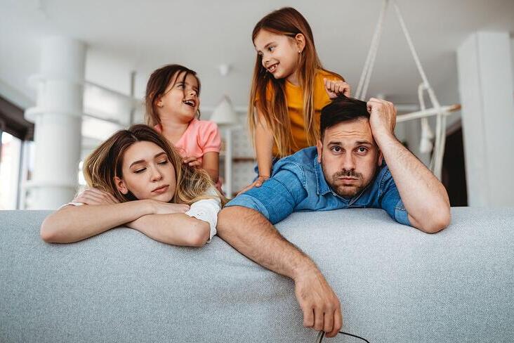 Exhausted parents leaning on a sofa with children playing behind them.