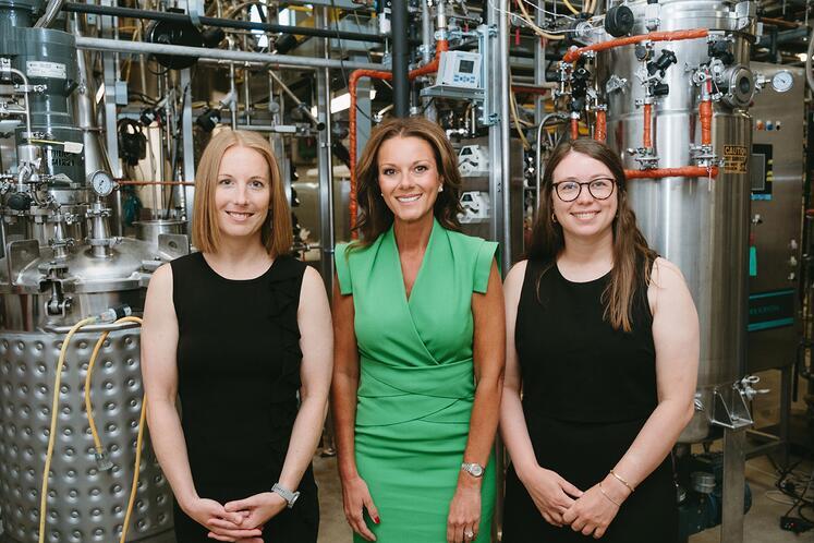 Three women standing in a bioprocessing laboratory with large stainless steel industrial equipment in the background.