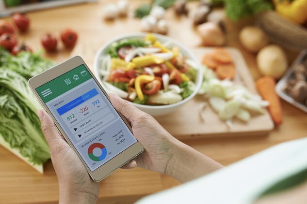 An individual’s hands holding a smartphone displaying food analysis information, on a backdrop of healthy food on a kitchen counter.