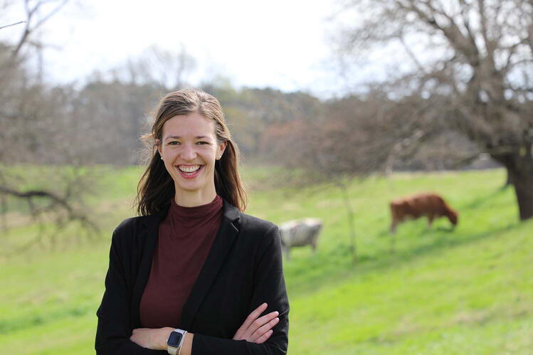 A smiling woman in a dark suit jacket stands with her arms crossed in front of a field with two cows shown in a blurred background