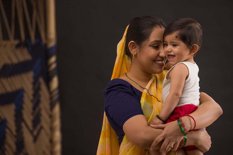 A woman in a yellow sari holds a smiling baby dressed in white and red.