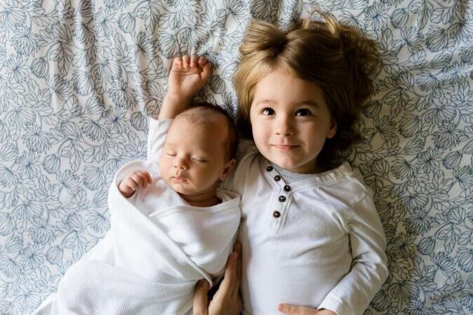 A toddler lies beside her infant sibling and smiles