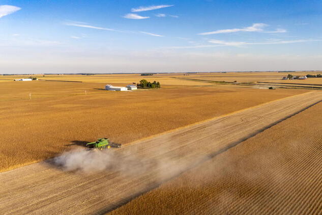 A combine harvester moves through a large, golden field under a blue sky; farm buildings are visible in the background