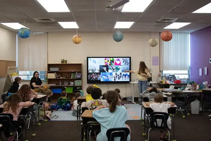 A person points at a screen with colorful images at the front of an elementary school classroom