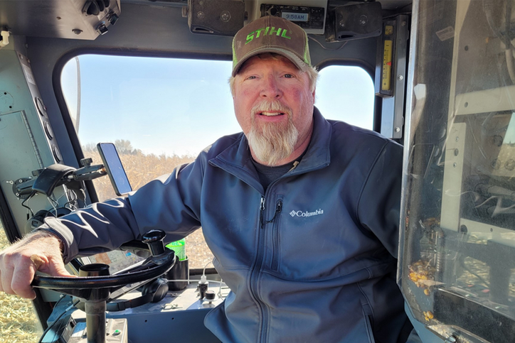 A bearded man wearing a ballcap sitting in a tractor with a field in the background