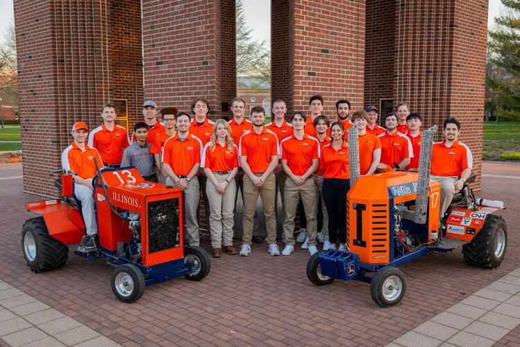 A large group of Illini Pullers students wearing matching team polos pose outdoors with two competition tractors.