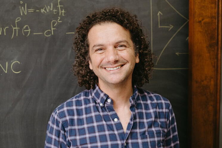 A man in a checkered shirt stands in front of a chalkboard with formulas written on it