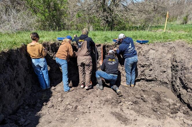 Five people work in a large trench, examining the soil layers. They are wearing casual outdoor clothing and hats. Trees and grass are in the background.