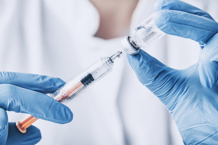 A close-up of a hands wearing surgical gloves holding an injection needle and a vial with liquid.