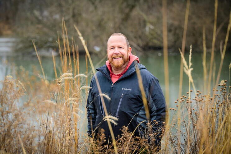 A man stands outside with tall grasses in the foreground and a pond in the background