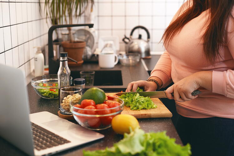 A woman chopping vegetables in a kitchen with a laptop on the counter